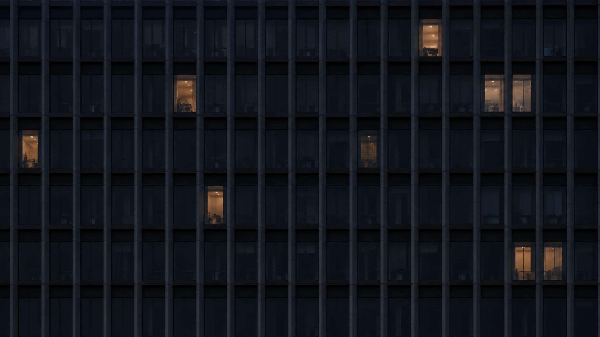 The window grid of an office façade at night, a few windows lit.